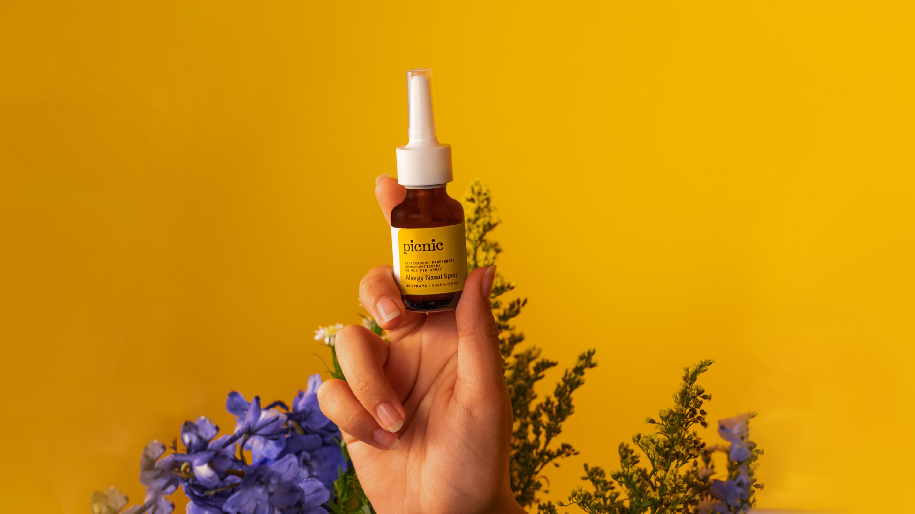 A person's hand holding up Picnic's nasal spray in front of flowers.