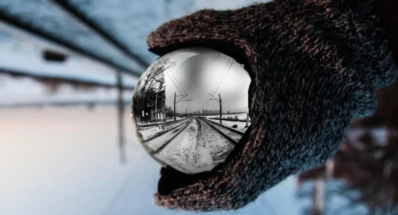 A gloved hand holding a reflective metal sphere showing train tracks in the snow.