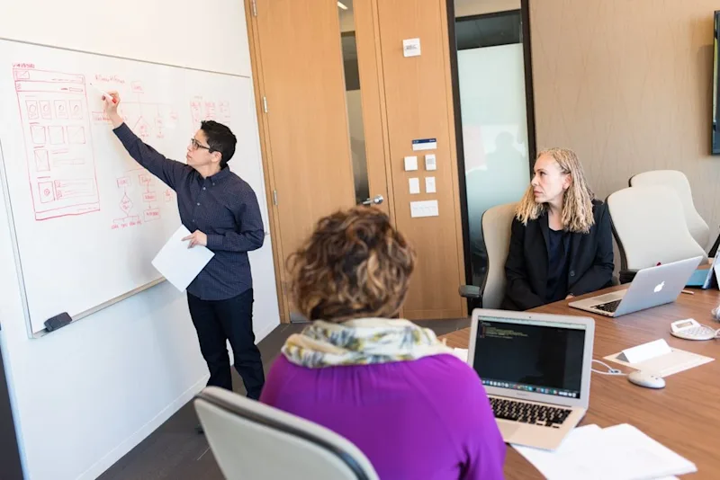 Three colleagues working in a conference room at an office. One is writing on a whiteboard while the other 2 sit at a table talking.