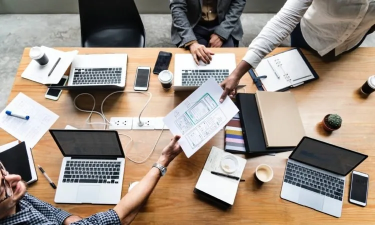 A top down view of 3 coworkers on laptops at a conference table and one of them is handing a piece of paper to the other.