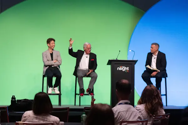 Will Jung, Jim Marous, and John Fimbel (left to right) sit on stools for a panel discussion at nCino nSight 2026, with Marous gesturing mid-conversation. A green and blue circular backdrop is behind them, and audience members watch from tables in the foreground.