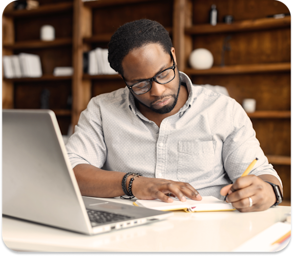 A man writes on a piece of paper with his laptop open on the desk.