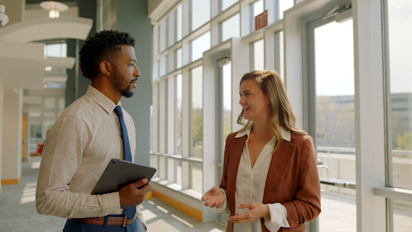 Two professionals talking in a sunlit office hallway — a man in a beige shirt and blue tie holding a tablet, and a woman in a rust blazer gesturing as she speaks.Two professionals talking in a sunlit office hallway