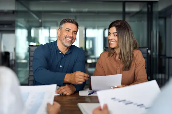 A man and woman sit side by side at a wooden desk in a glass-walled office, smiling as they review paperwork with someone seated across from them.
