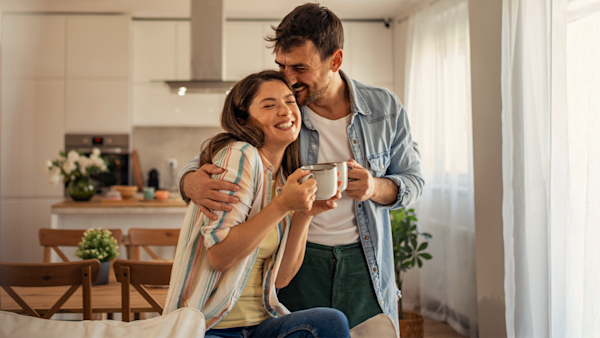 A smiling couple embraces in a bright, modern kitchen while holding a shared mug of coffee, with a dining table, potted plants, and soft natural light in the background.