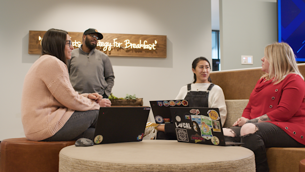 Four people sit and stand around a laptop table at nCino's modern office setting, engaged in discussion with a wooden sign in the background.
