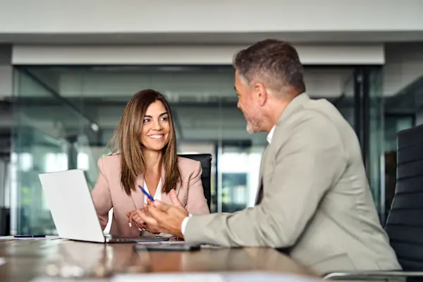 Two business professionals talking over a laptop at a desk in a modern office.