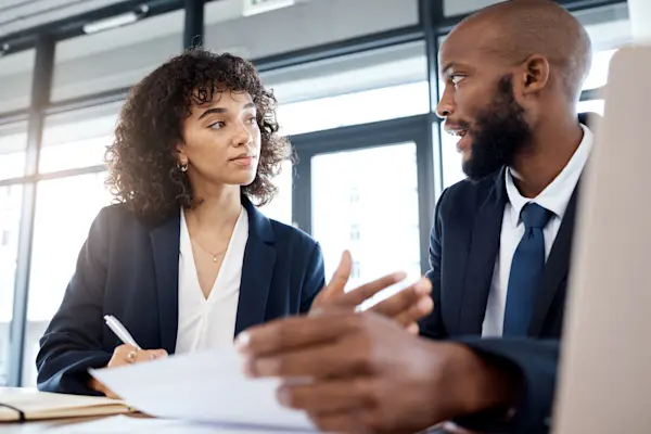 Two banking professionals in a discussion at an office desk, one taking notes.