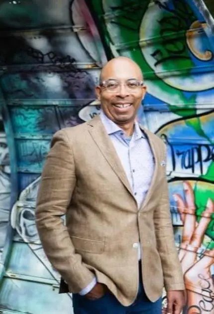 A man with glasses in a tan jacket and blue pants smiles in front of a wall covered in colorful graffiti.