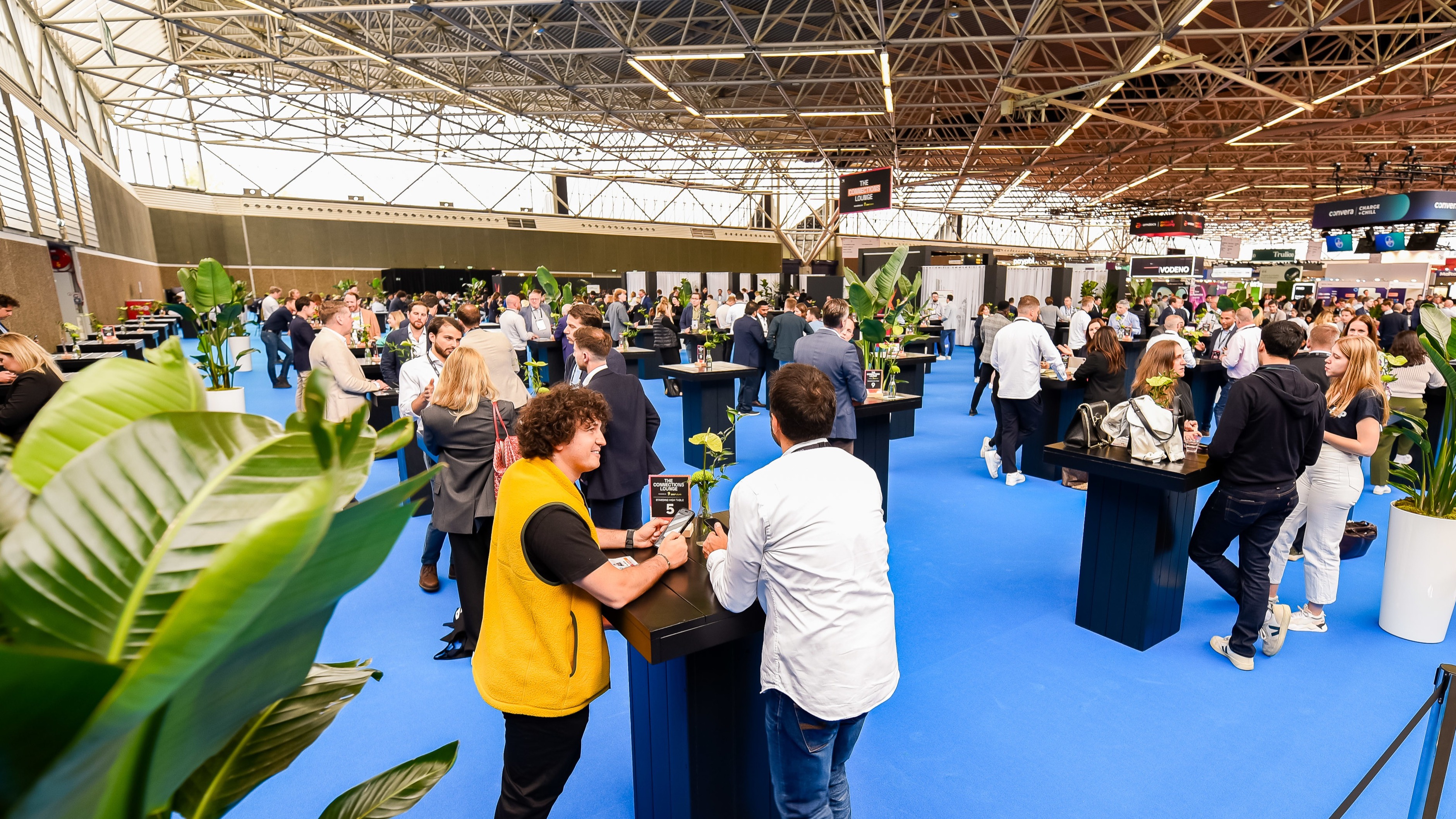 A wide-angle shot of the show floor featuring a large blue carpet, plants, and tables with professional-looking people standing around, talking, and networking.