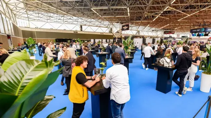 A wide-angle shot of the show floor featuring a large blue carpet, plants, and tables with professional-looking people standing around, talking, and networking.