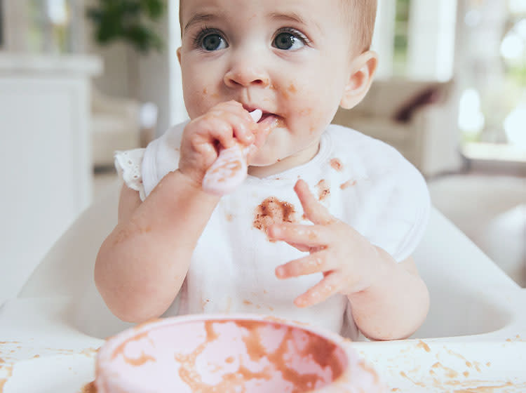A child with food stains on his face and clothes sits next to the table, holding a spoon in his hands