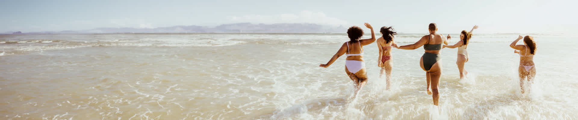 Quattro donne corrono felici tra le onde dell'oceano in una giornata di sole, godendosi una divertente giornata in spiaggia.