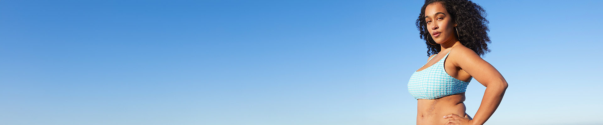 Una donna con i capelli ricci sta sulla spiaggia indossando un bikini a quadretti blu, sullo sfondo si possono vedere le onde del mare.