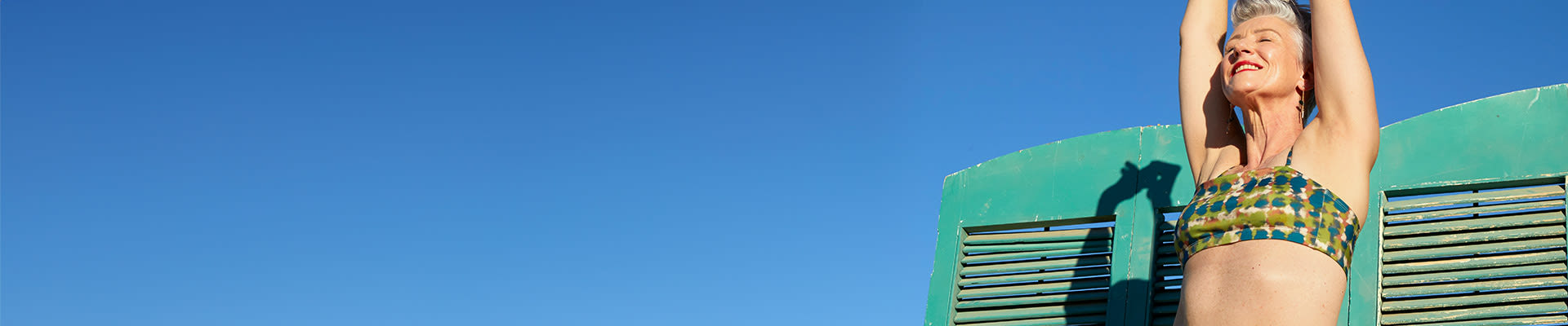 Una donna matura tiene le mani alzate alla luce del sole, con gli occhi chiusi, appoggiata a una porta di legno turchese sulla spiaggia