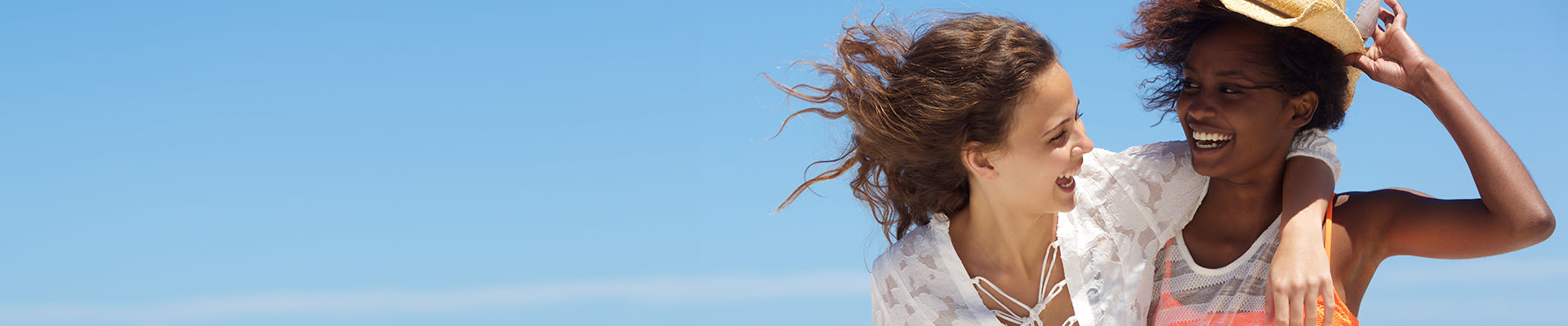 Due giovani donne sorridono con gioia e si abbracciano sulla spiaggia con un cielo azzurro sullo sfondo.