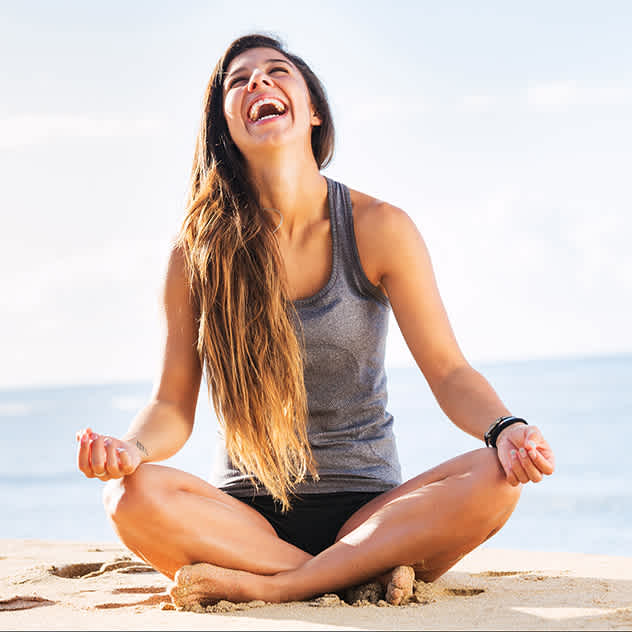 Una donna con i capelli lunghi è seduta a gambe incrociate sulla spiaggia e ride gioiosamente con l'oceano sullo sfondo.