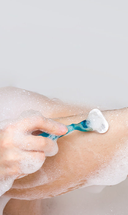 Una persona che usa un rasoio per radersi la gamba con sapone e bolle su uno sfondo chiaro.