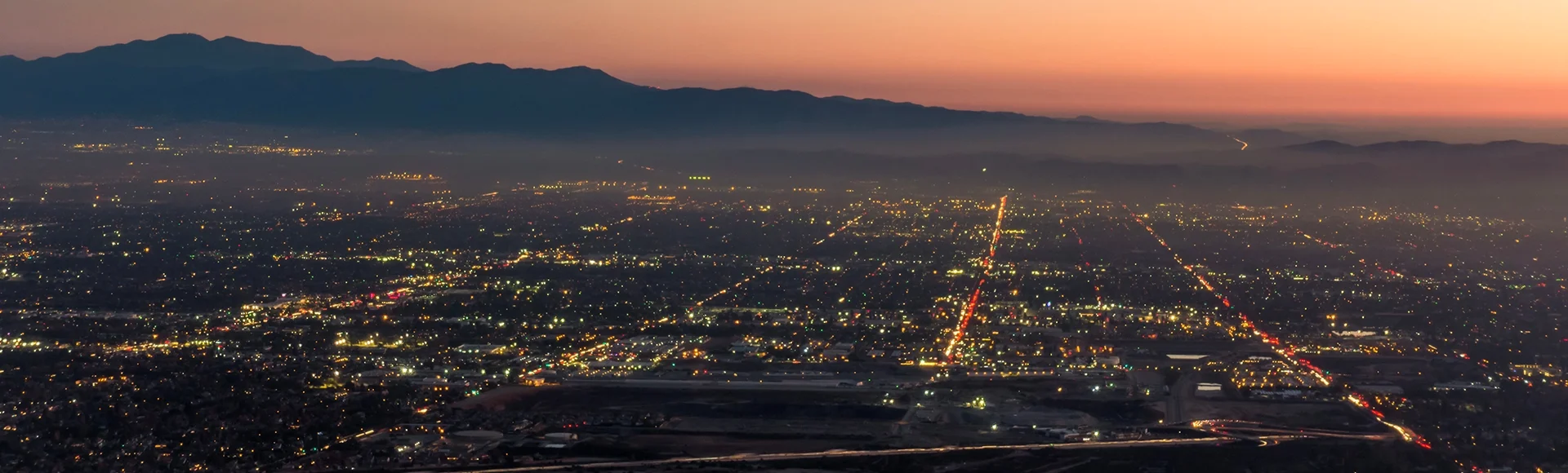 The city lights of the skyline of the Inland Empire near Los Angeles California begin to appear as the sun sets in a dramatic orange sunset. View from Potato Mountain in Claremont Wilderness Par