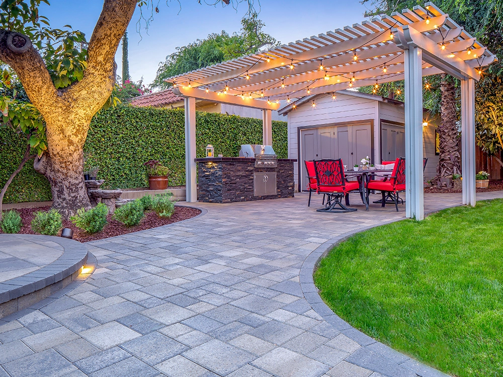 Landscaped backyard with pergola over an outdoor kitchen BBQ and paving stone patio. Also includes installed landscape lighting. 