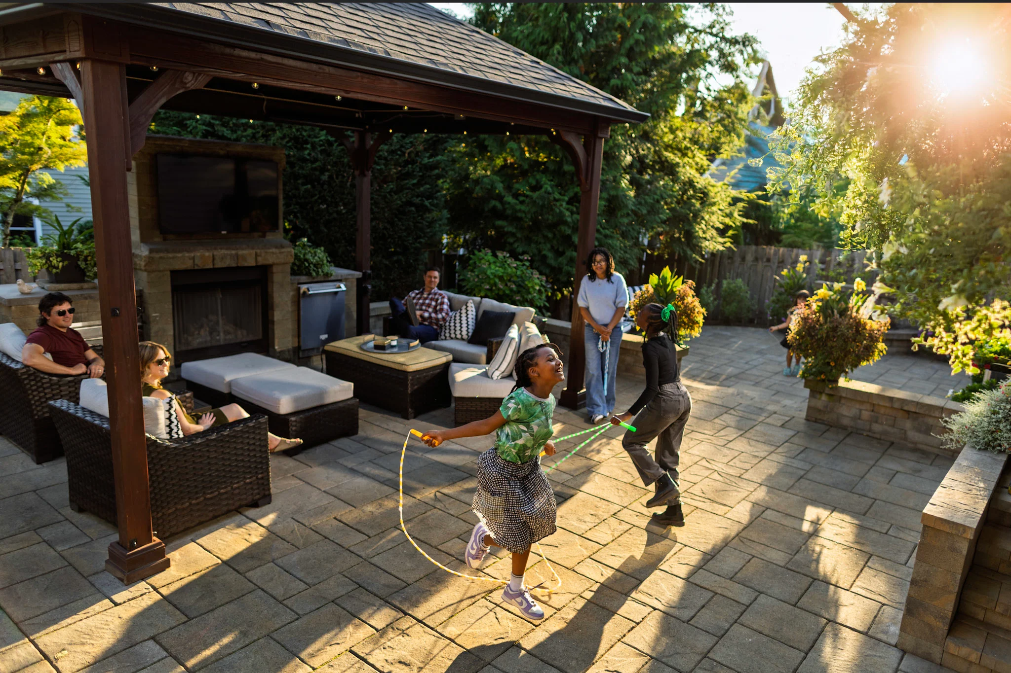 Family and friends on paver patio under pergola as children play 