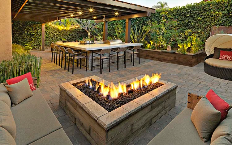 Fire pit, gas fire pit, lit, L-shaped fire pit, outdoor lighting, pergola, outdoor kitchen, grilling station, countertop, total outdoor transformation, pavers, paver patio, patio furniture, retaining wall, paving stones, universal region, daytime, evening