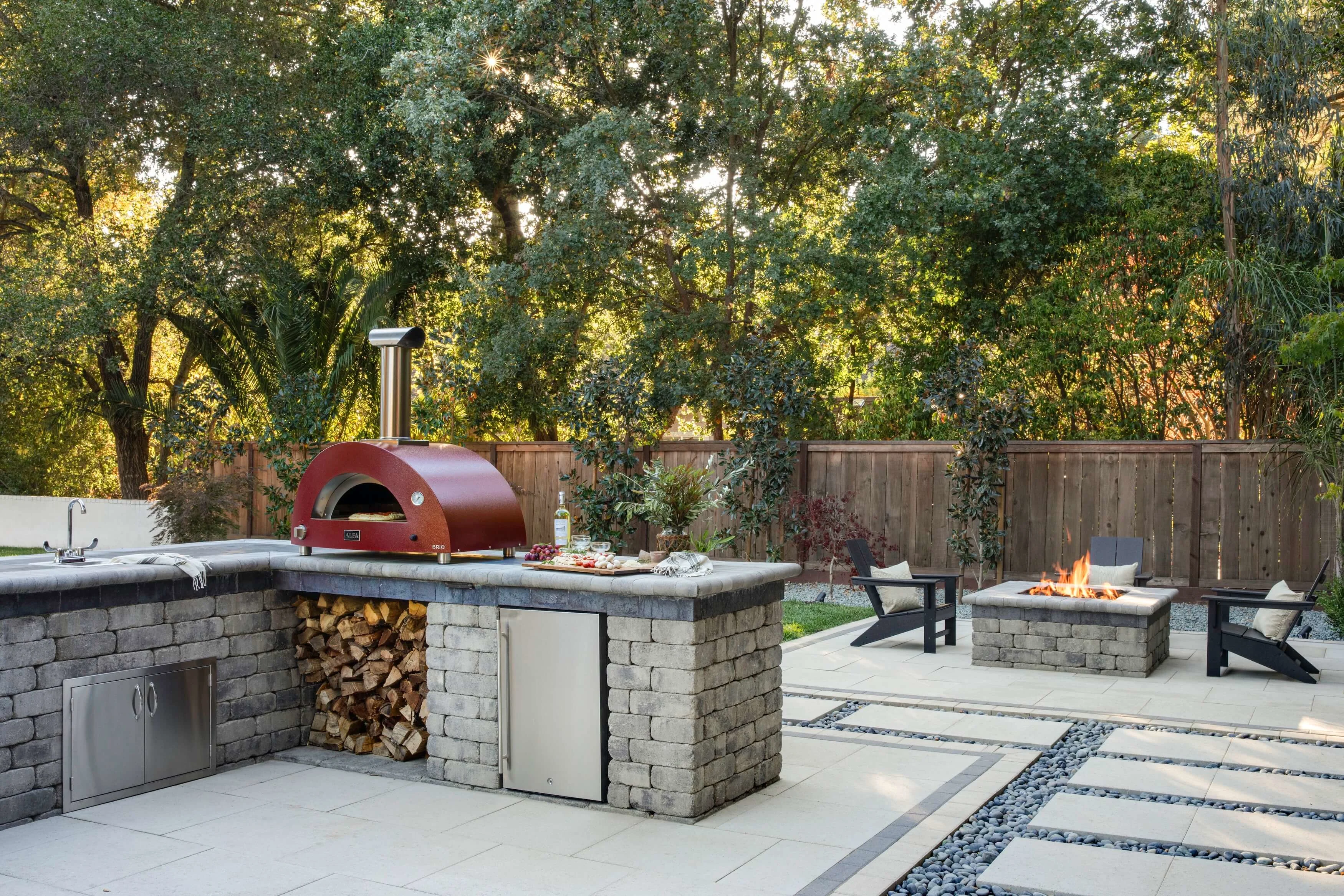 View of outdoor kitchen with pizza oven and modern square fit pit area in the background
