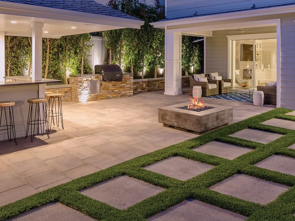 A lovey paver patio with turf insets and built-in fire pit at night.