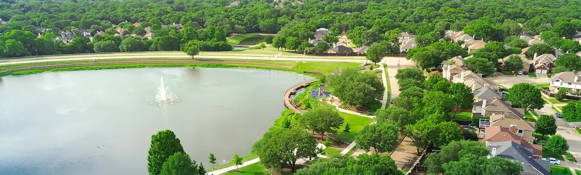 Lush green community Rheudasil Park in Flower Mound, Texas with curved boardwalk, walking trails, playground, upscale suburban two-story houses with resort-style swimming pool, grassy lawn, aerial