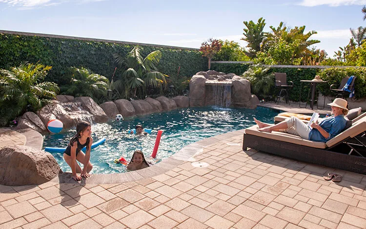 Three children swimming in a pool surrounded by paver pool deck while older white woman watches