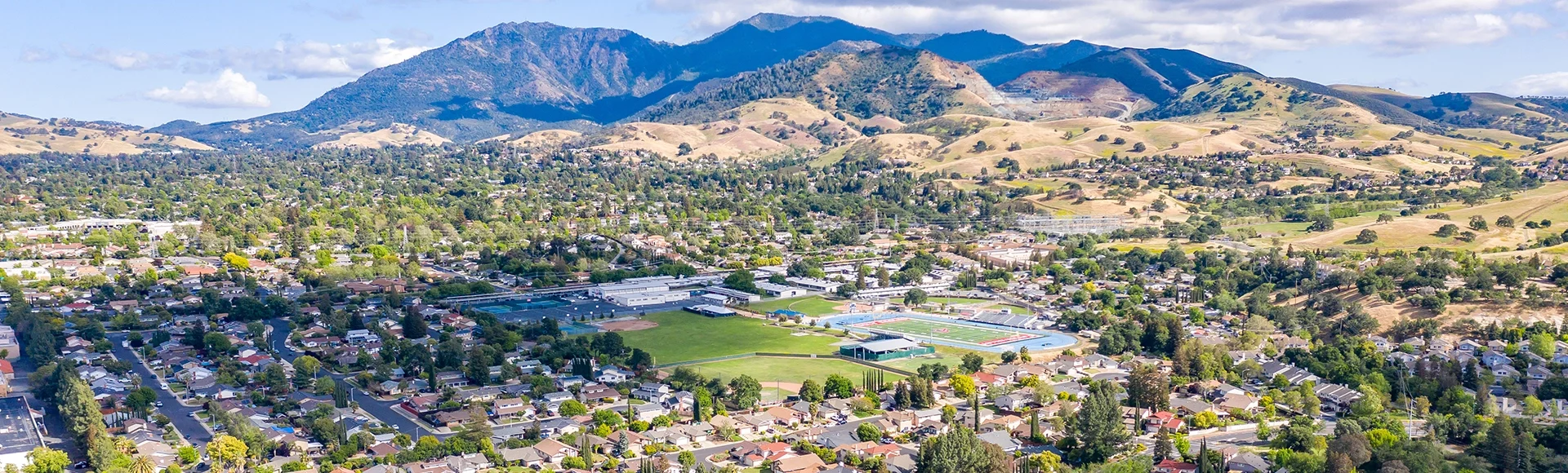 Aerial drone view of Concord, California, featuring residential neighborhoods, a sports complex, and Mount Diablo in the scenic background under a partly cloudy blue sky.