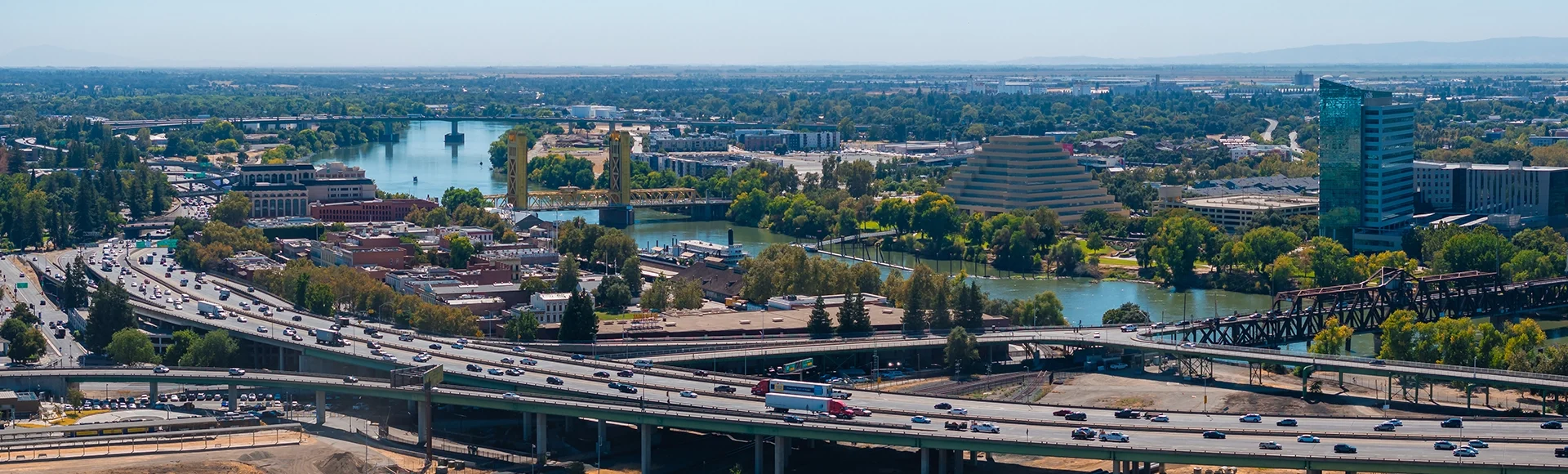 
Aerial view of Sacramento, California, featuring the yellow gold Tower Bridge, the Ziggurat building, highways, and lush greenery under a clear sky.
