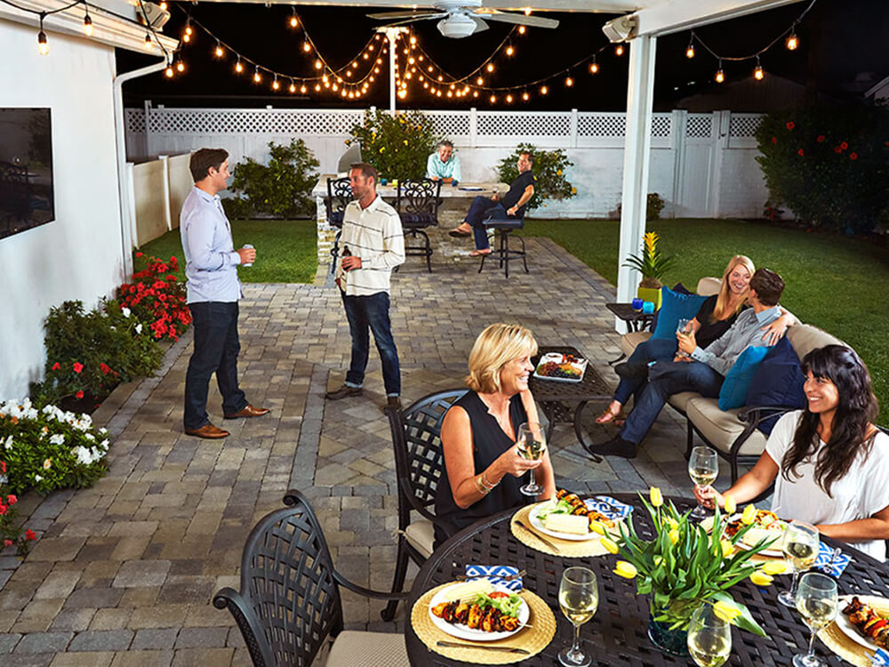 Stone patio with pergola with various adults, some having dinner and chatting. 