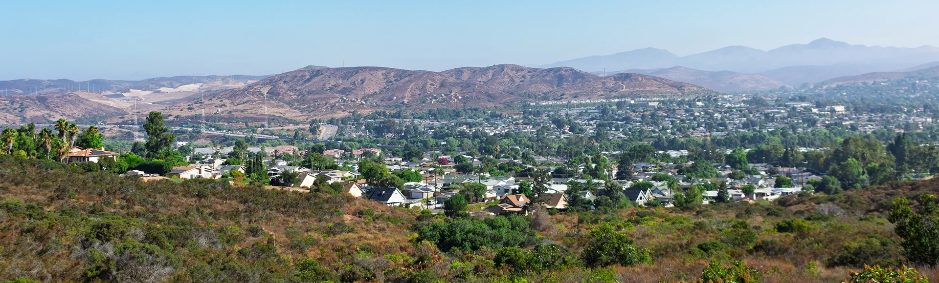 A scenic view from Big Rock Trail overlooking Santee, California, nestled among arid hills and natural vegetation, with distant mountains under a clear blue sky. 