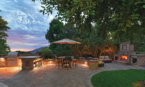 Outdoor fireplace, pavers, outdoor kitchen, BBQ, grilling station, outdoor lighting, retaining wall, artificial turf, cozy, fire, lit, paver patio, patio furniture, outdoor living, total outdoor transformation, backyard, fire, paving stones, sunset, universal region, evening