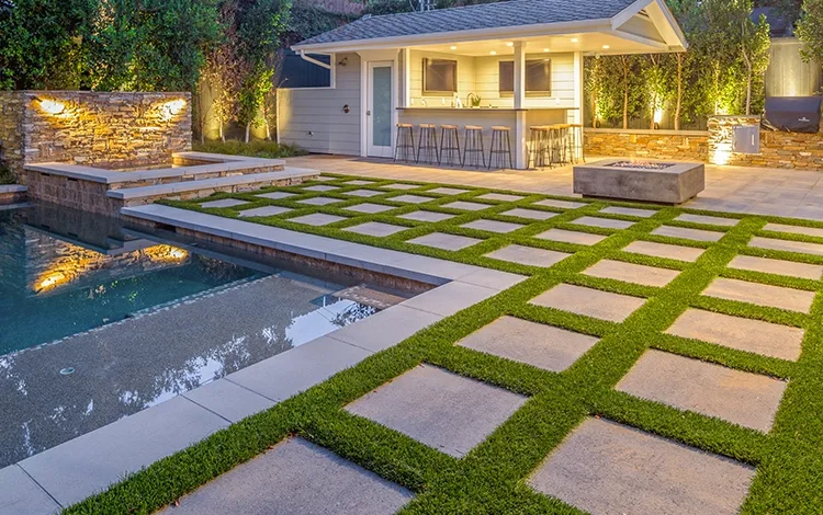 Large patio with large paving stones, artificial turf around a pool with a stand-alone outdoor kitchen and paving stone fire pit. 