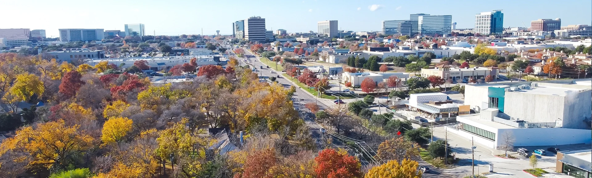 Yellow red orange fall foliage over urban park along Belt Line Road with office buildings, skylines, commercial complex in North Dallas and Addison, sunny clear blue sky, vibrant autumn leaves