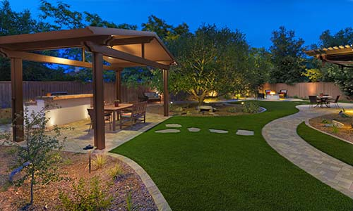 Artificial turf, backyard, pergola, outdoor kitchen, outdoor lighting, paver walkway, fire pit, outdoor seating, patio furniture, artificial grass, total backyard transformation, universal region, nighttime, evening