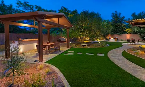 Artificial turf, backyard, pergola, outdoor kitchen, outdoor lighting, paver walkway, fire pit, outdoor seating, patio furniture, artificial grass, total backyard transformation, universal region, nighttime, evening