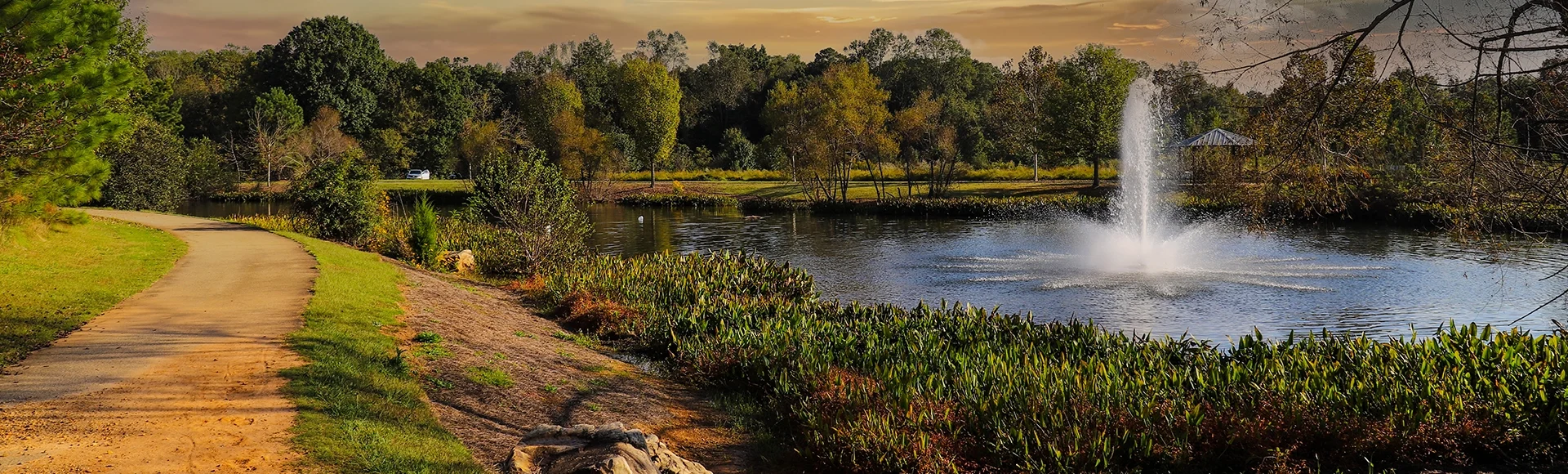 A shot of winding footpath along a lake with a water fountain surrounded by lush green and autumn colored trees and plants with powerful clouds at sunset at Garrard Landing Park in Alpharetta