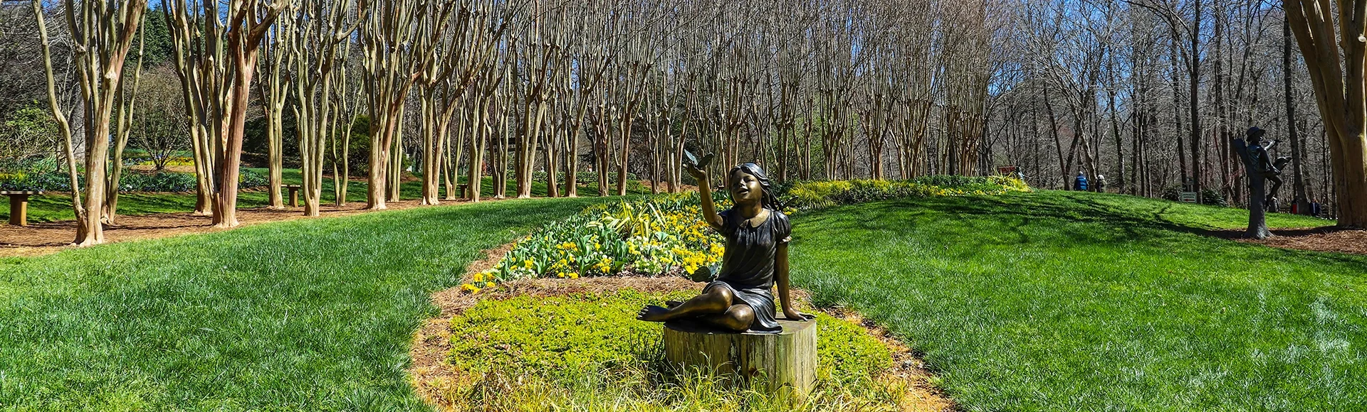 A copper statue of a little girl holding a butterfly surrounded by lush green grass, bare winter trees, colorful flowers and lush green plants with blue sky at Gibbs Gardens in Ball Ground Georgia USA 