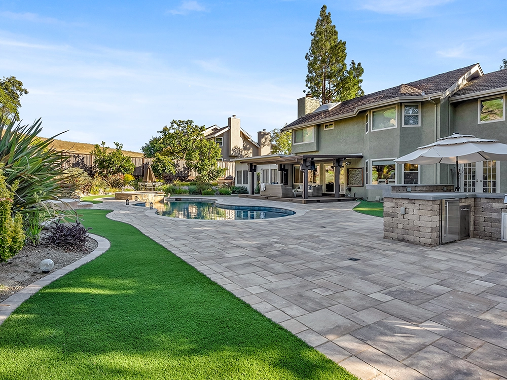 Large paver patio with artificial turf lawn by a pool with a custom System Pavers outdoor kitchen. 