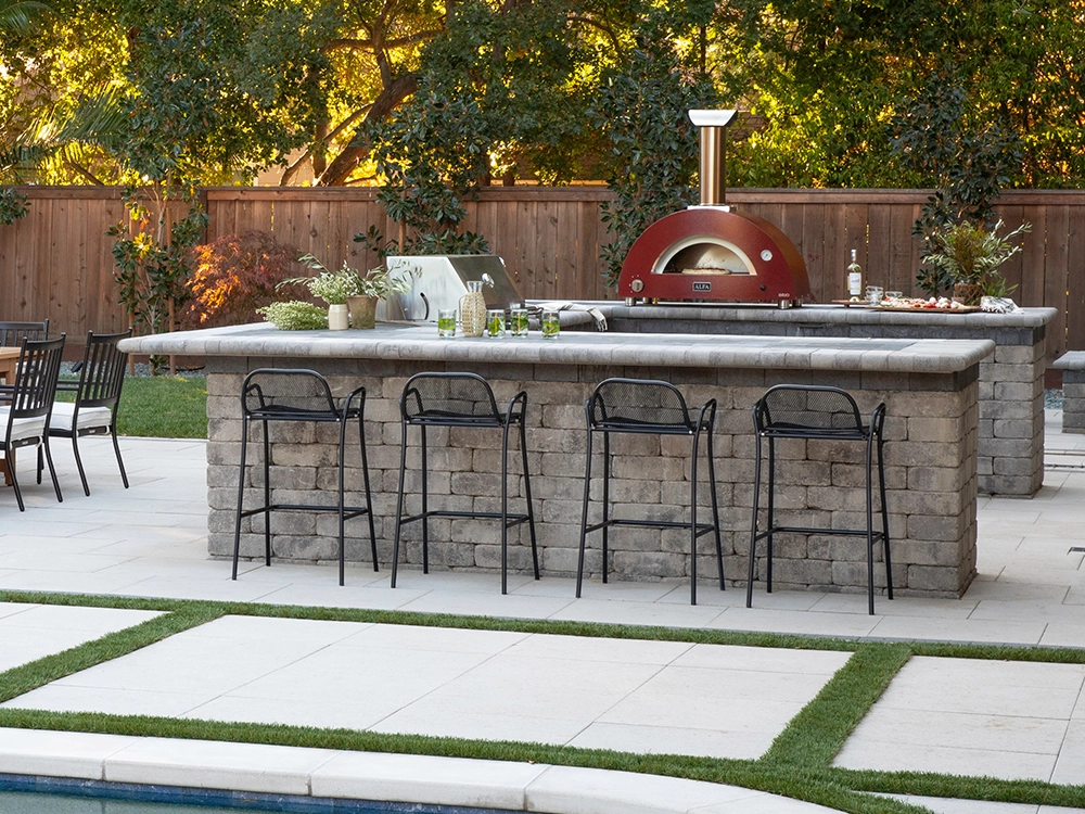 Large paving stone outdoor kitchen with bar stools and red pizza oven in a backyard. 
