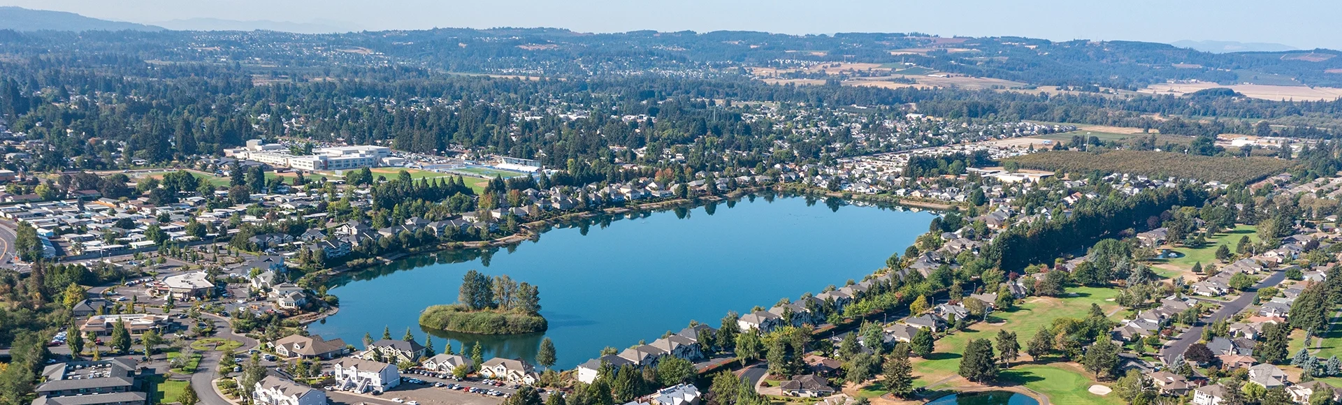 Aerial drone view of Staats Lake in Keizer, Oregon, near Salem, featuring a residential neighborhood, a golf course, and lush green landscapes under a clear blue sky