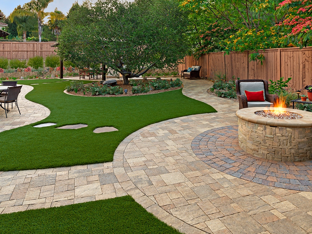 Large backyard with turf and paver walkways around a beautiful stone fire pit. 