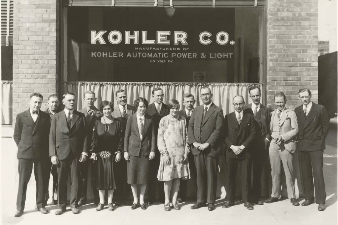 An old photograph with 15 men and women wearing executive clothes and standing in front of a Kohler office. 