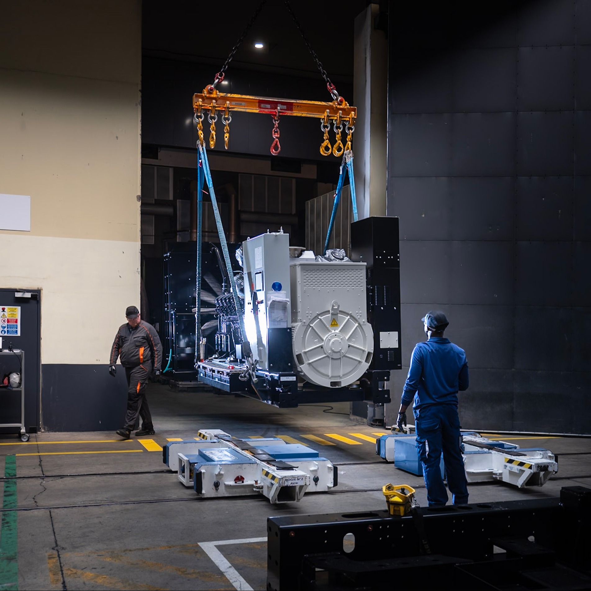 Industrial generator being crane-lifted during factory testing and commissioning in a manufacturing facility.
