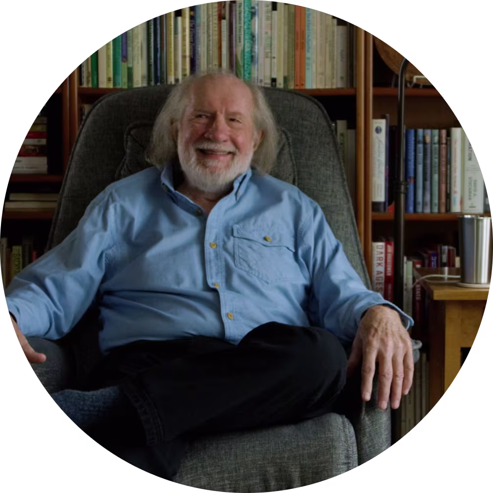 Elderly man wearing a blue shirt and dark pants, comfortably sitting on a grey chair in a home library.