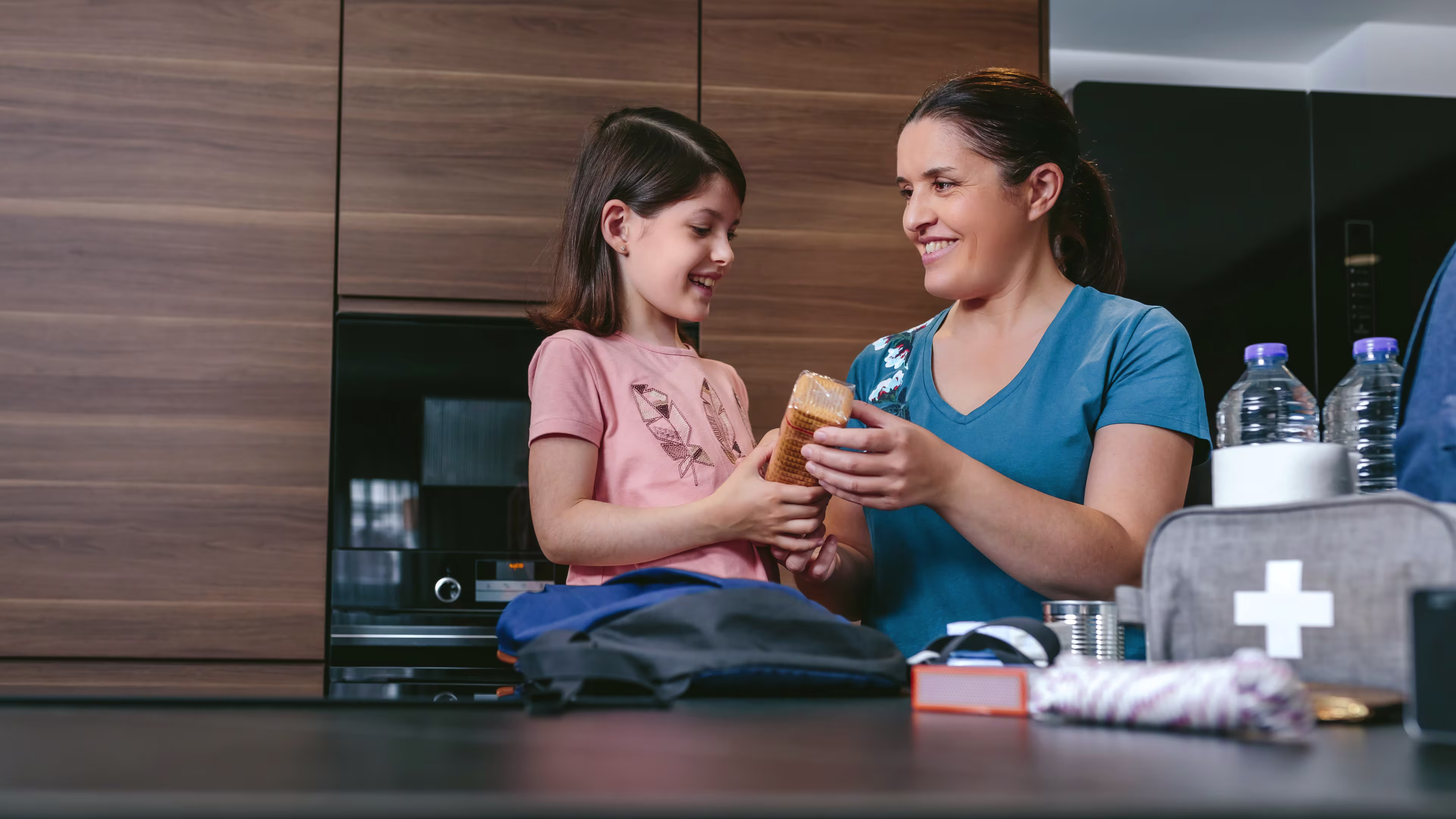 A cheerful mother and daughter in a bright kitchen, packing a backpack together with snacks and emergency supplies.