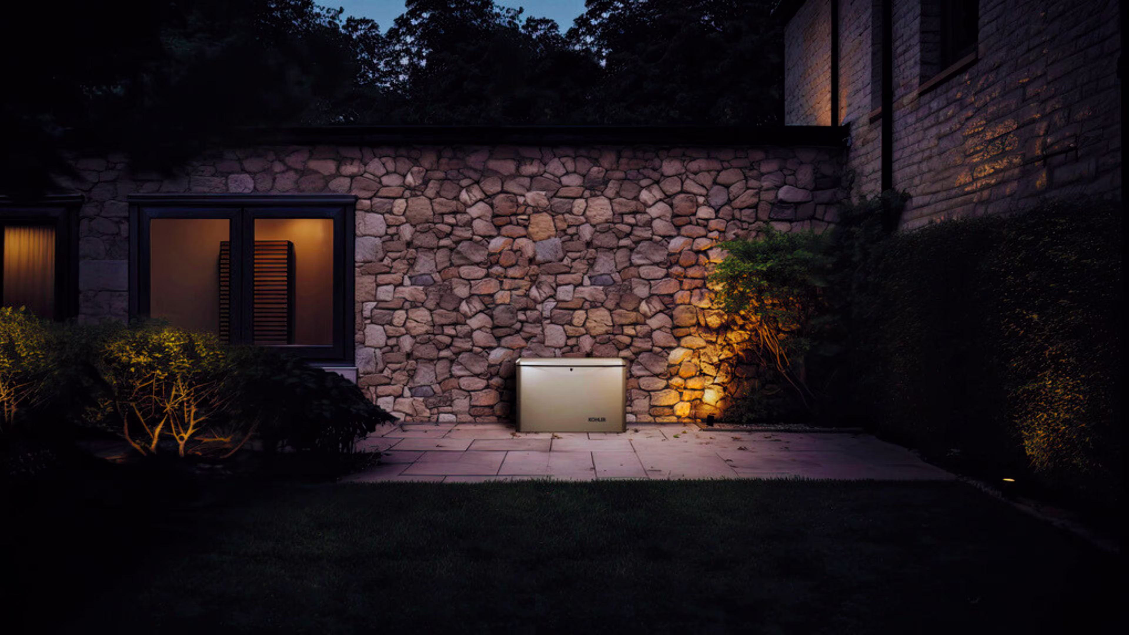 A KOHLER generator sitting in the yard of a stone house in a dark scenario illuminated by lights